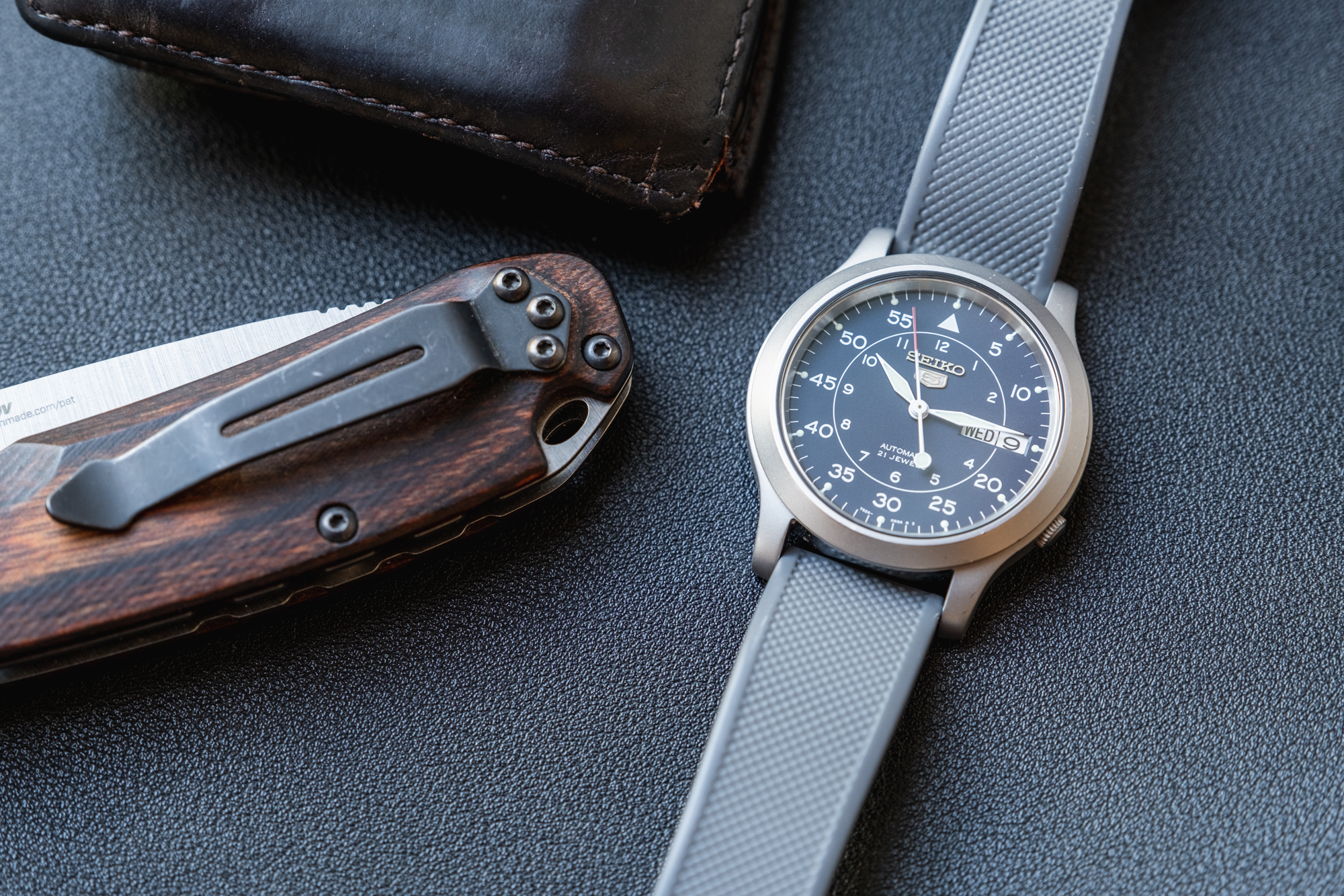 Close up of a blue Seiko 5 watch with a gray band next to a knife and a wallet