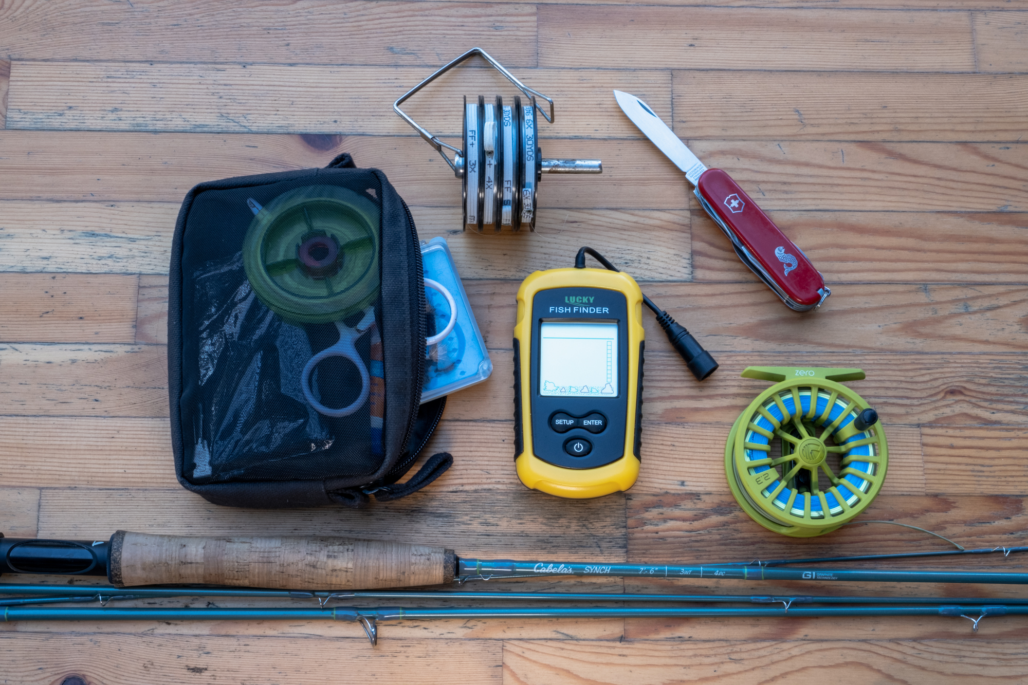 An assortment of cheap fishing gear on a table including swiss army knife, fly rod, reel, and a fish finder
