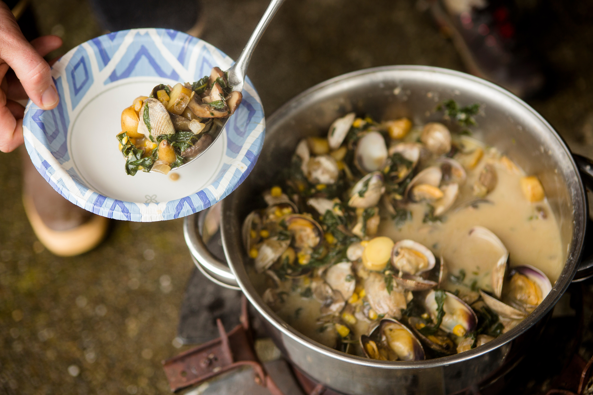 A metal spoon serving steaming clams, greens, and broth into a bowl from a large metal pot