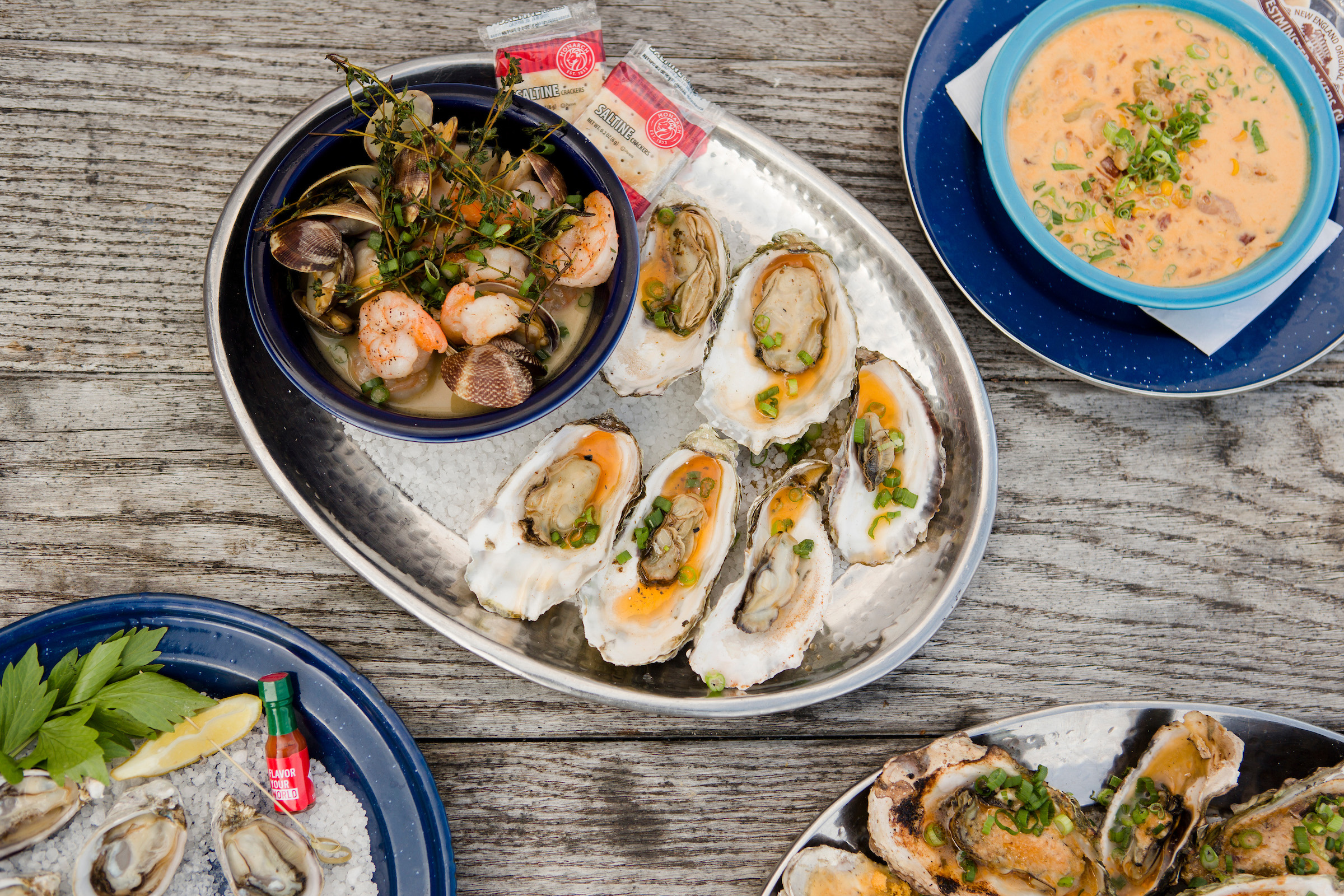 A large plate of oysters on a bed of salt with a bowl of shrimp and clams surrounded by plates of corn chowder and oysters