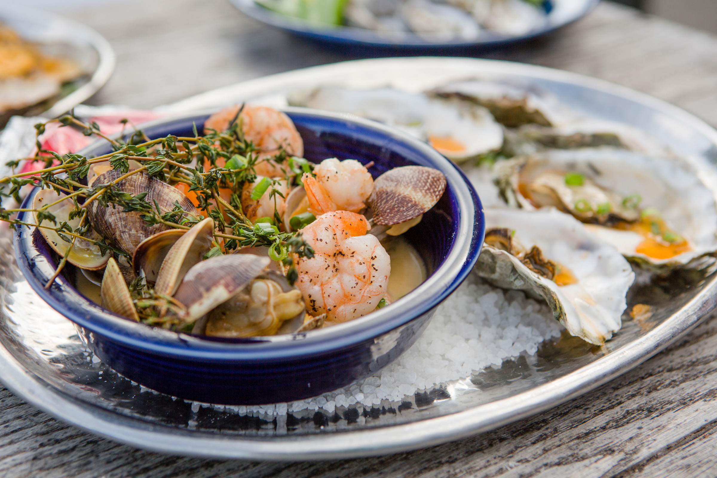 Steamed clams and cooked shrimp in a bowl topped with thyme next to a plate of oysters on a bed of salt