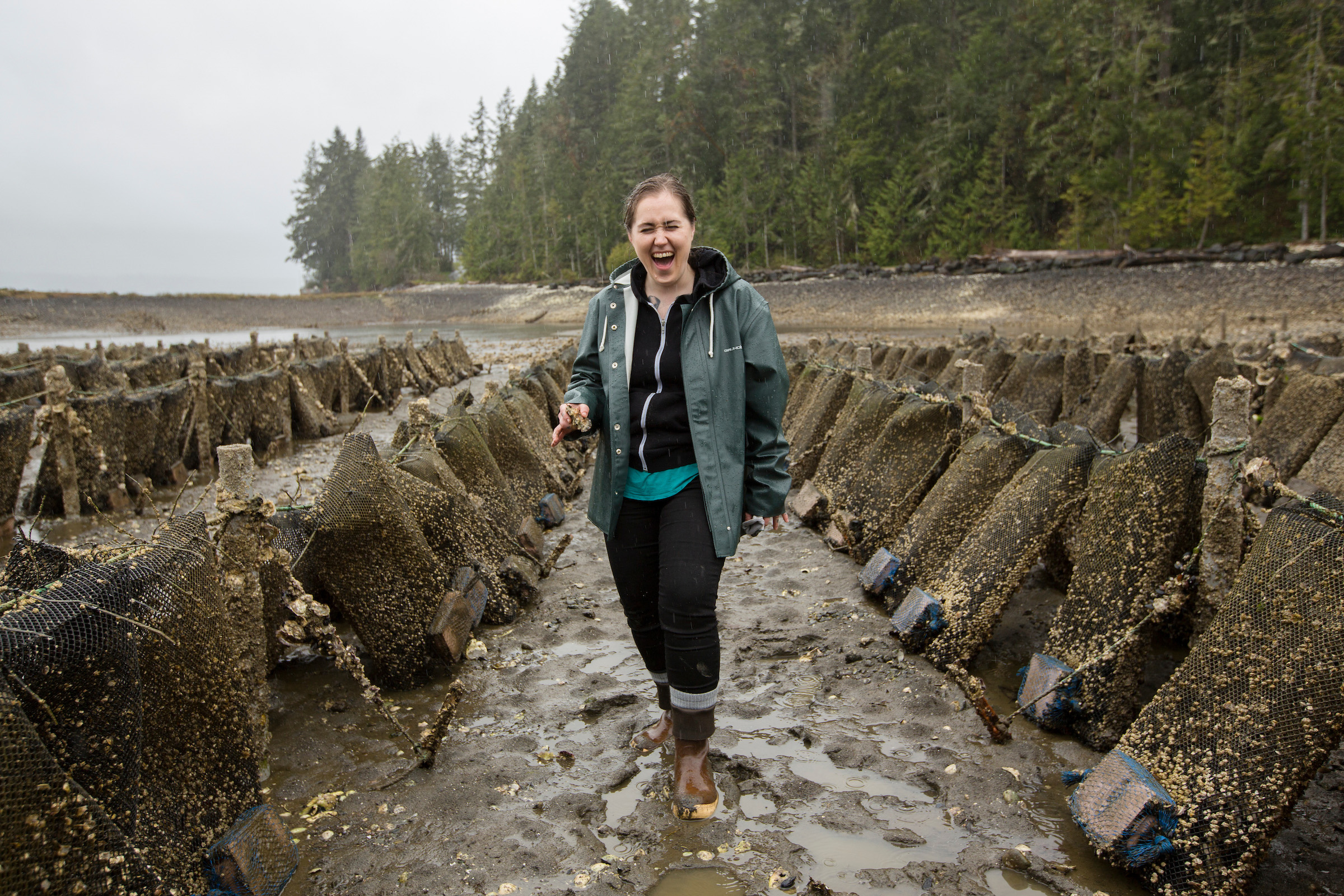 Chef Sara Harvey standing in a row of bagged oysters laughing and holding an oyster