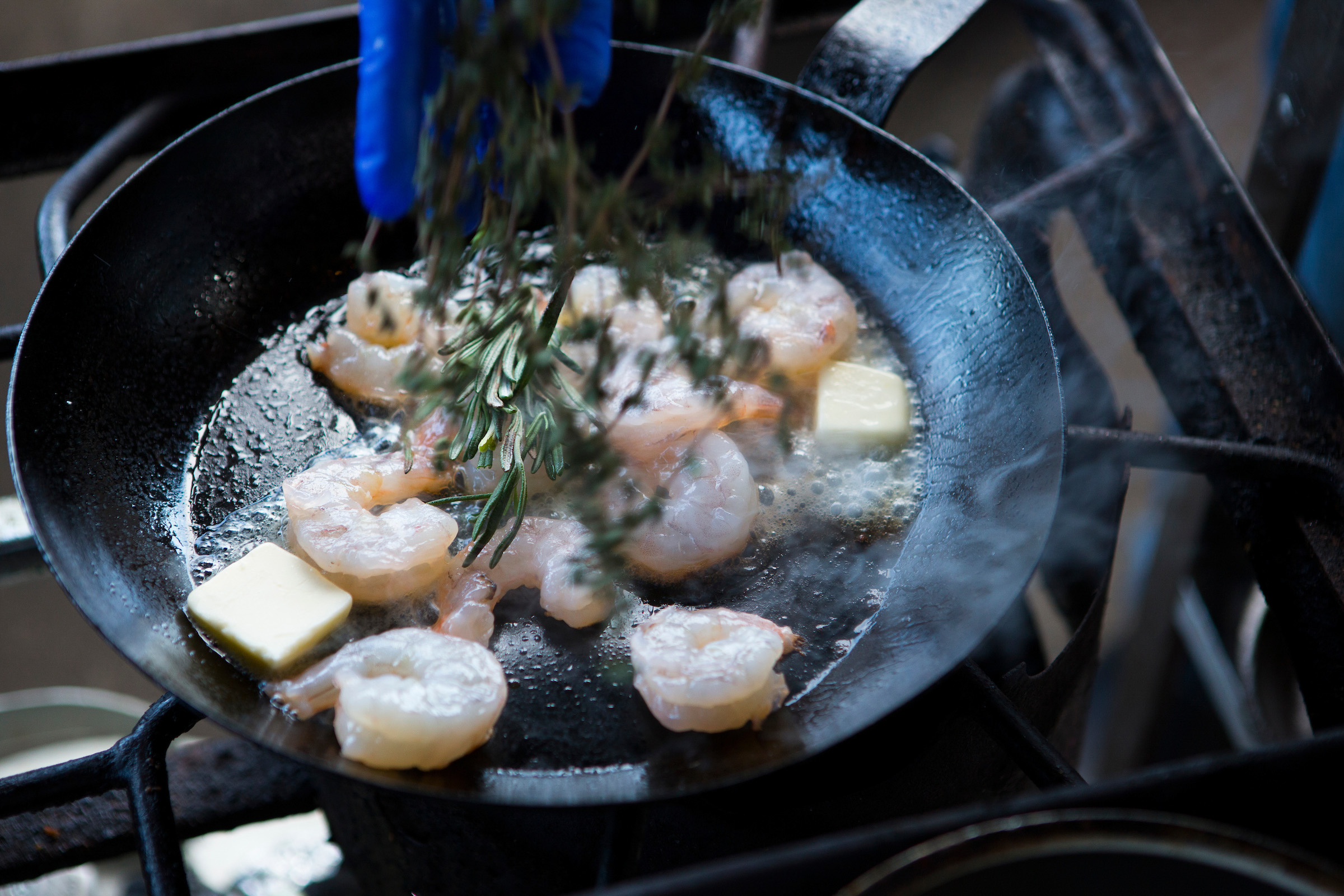 A closeup image of a chef dropping fresh herbs into a hot pan filled with button and raw shrimp