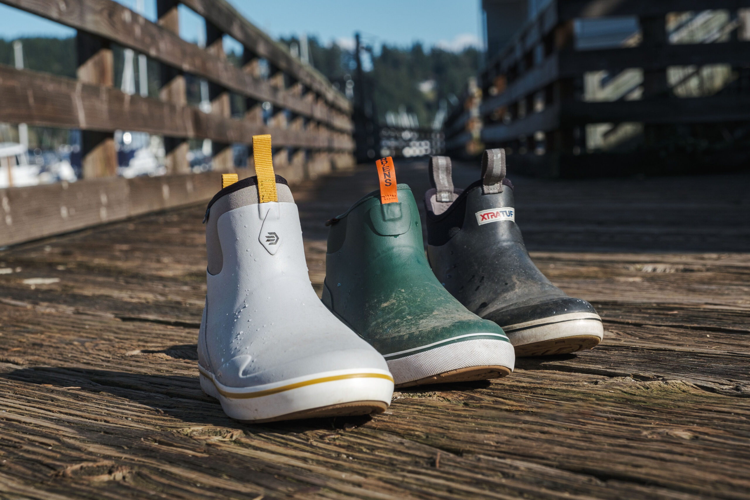 Three different deck boots from LaCrosse, Grundens, and Xtratuf lined up on a wood deck.