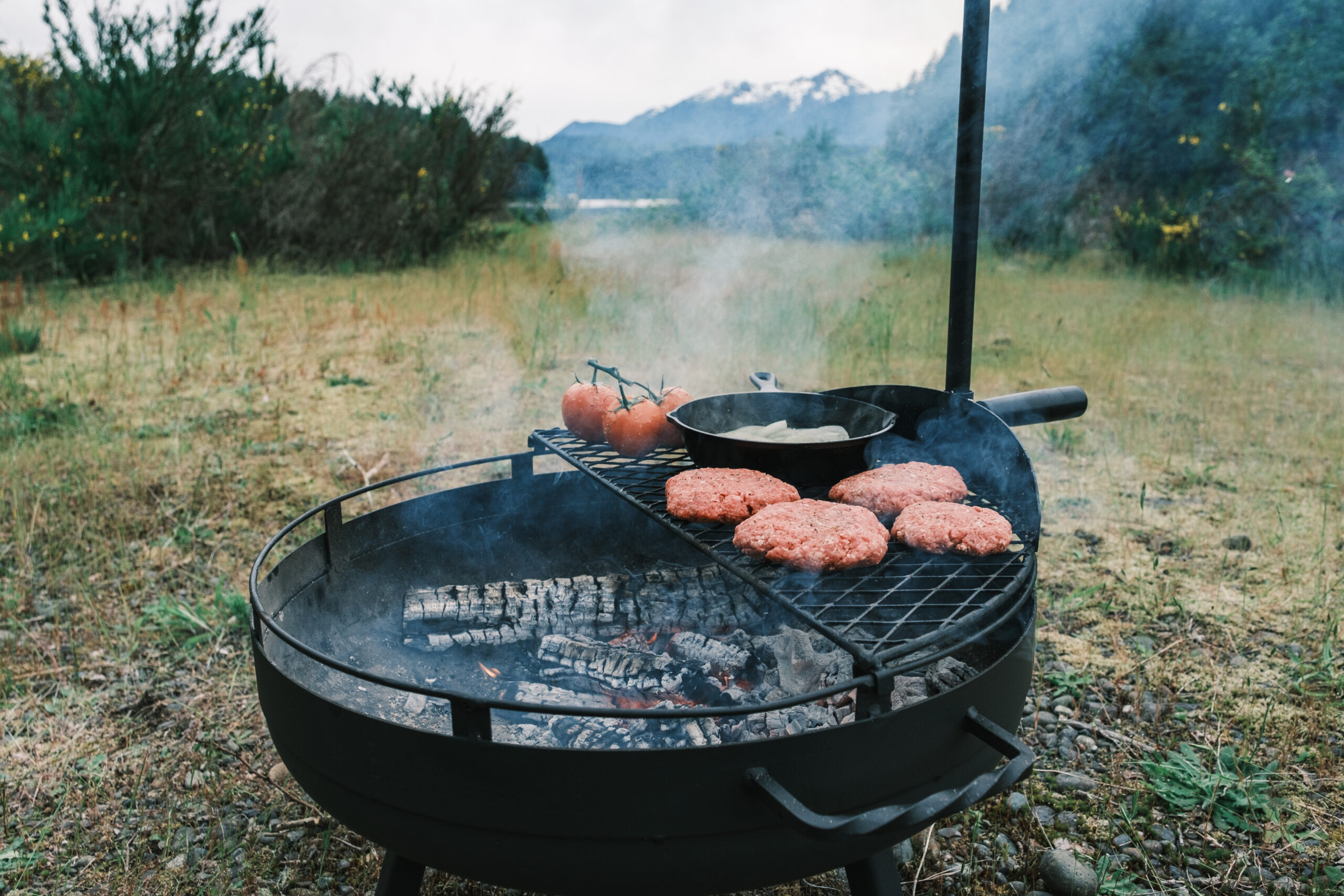 Burgers, tomatoes, and a black pan cooking over white wood coals in a black fire pit.