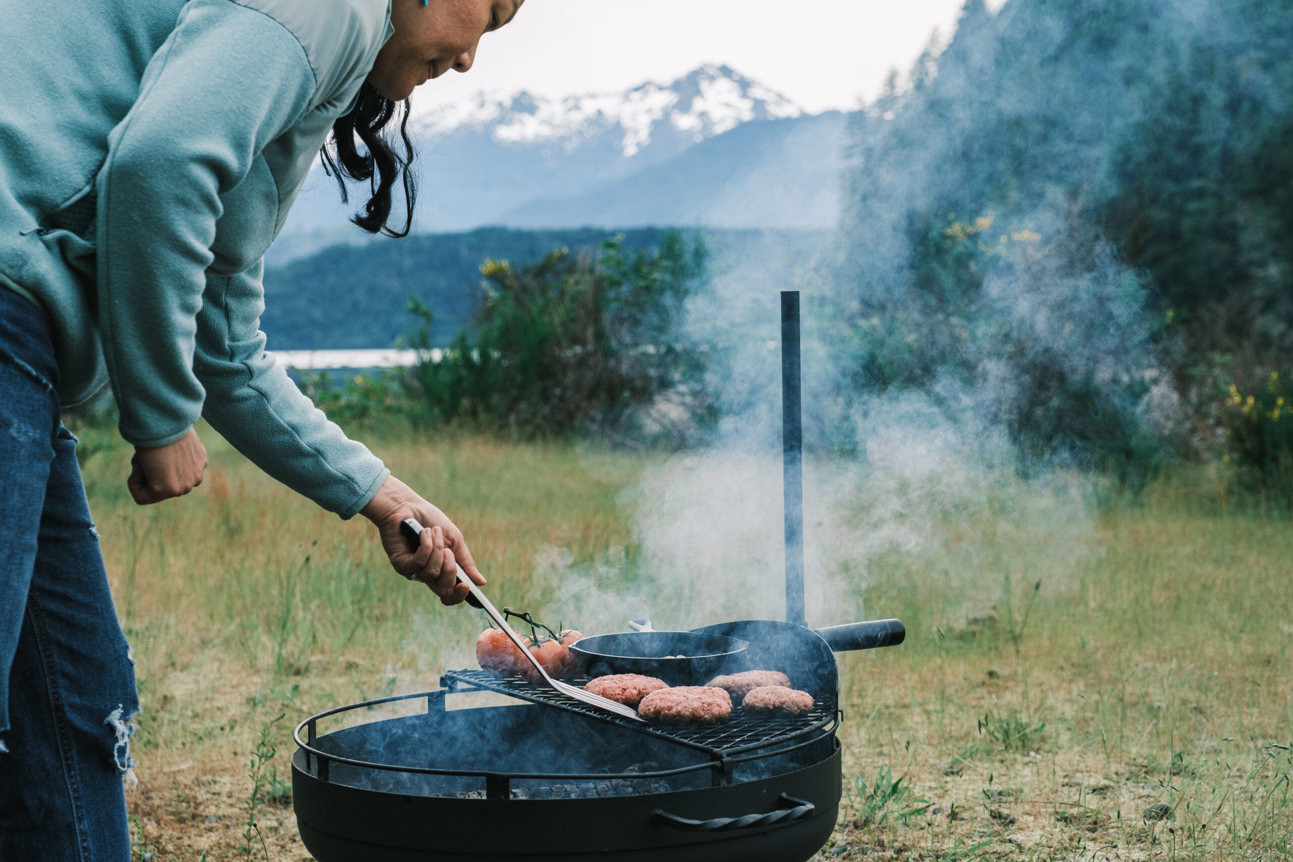 A woman holding a spatula and flipping burgers on a grill grate with mountains in the background